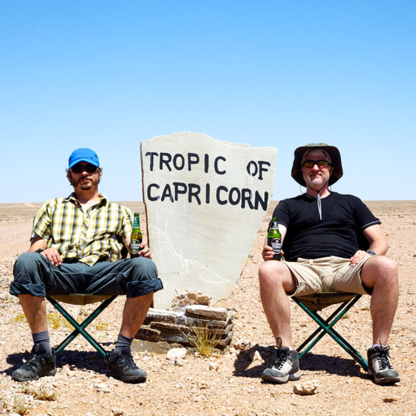 Two hikers sitting in chairs with a beer next to Tropic of Capricorn sign