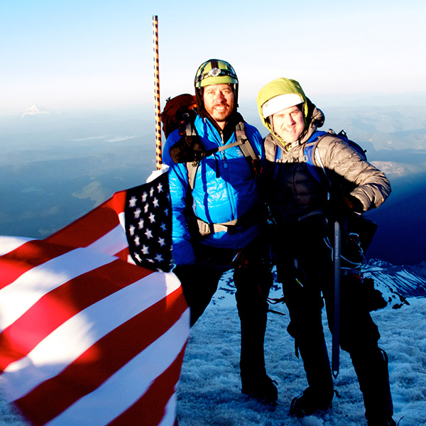 Two men standing atop Mount Hood with American flag